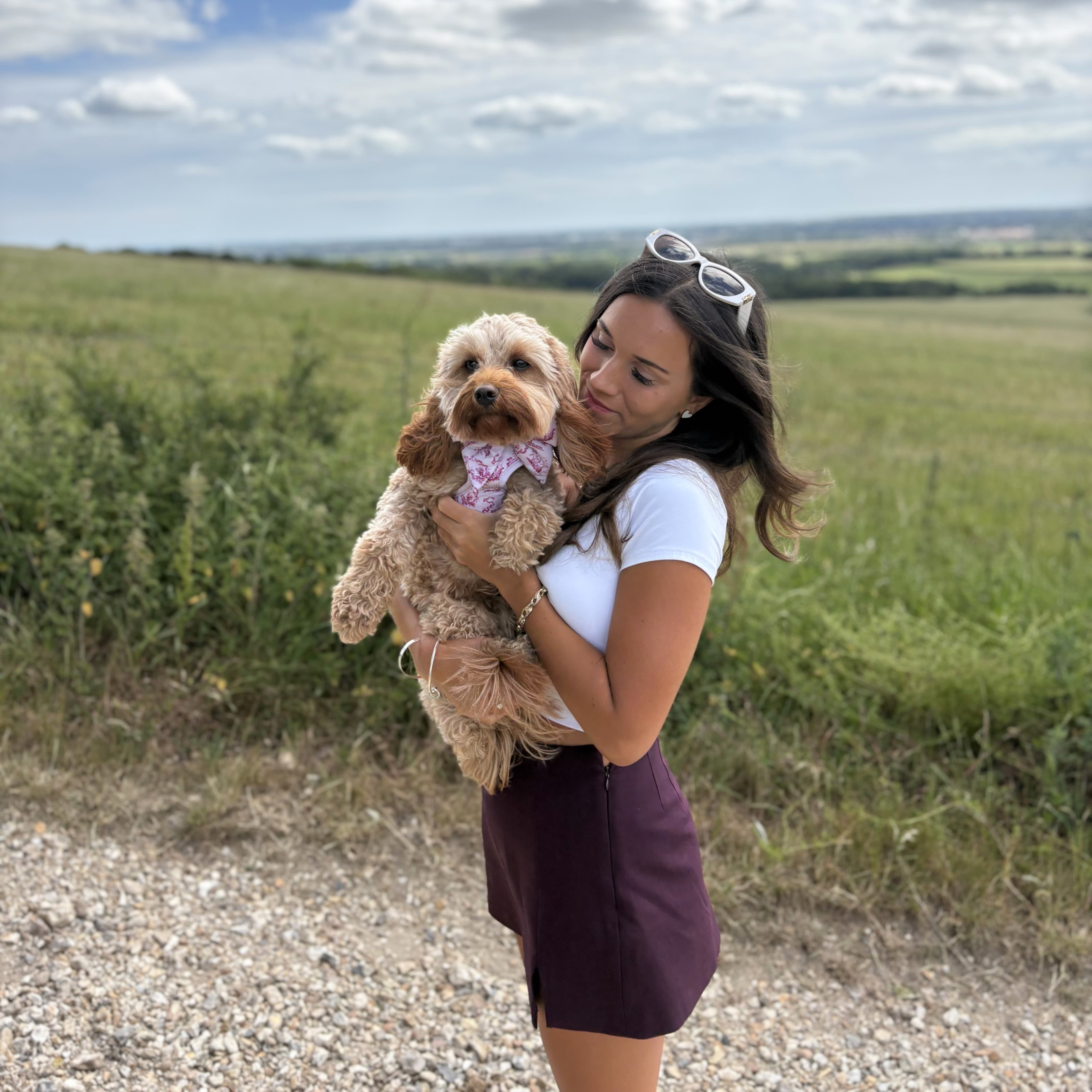 Woman holding small dog in floral bandana, enjoying scenic outdoor walk with luxury pet accessories.